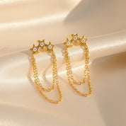 A pair of gold star-shaped dangle earrings with small clear stones and double chain drops, displayed on a cream satin fabric background.