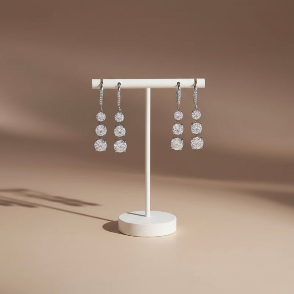 A pair of silver-toned dangle earrings with three clear round-cut stones each, displayed on a white jewelry stand against a warm beige studio background.