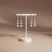A pair of silver-toned dangle earrings with three clear round-cut stones each, displayed on a white jewelry stand against a warm beige studio background.