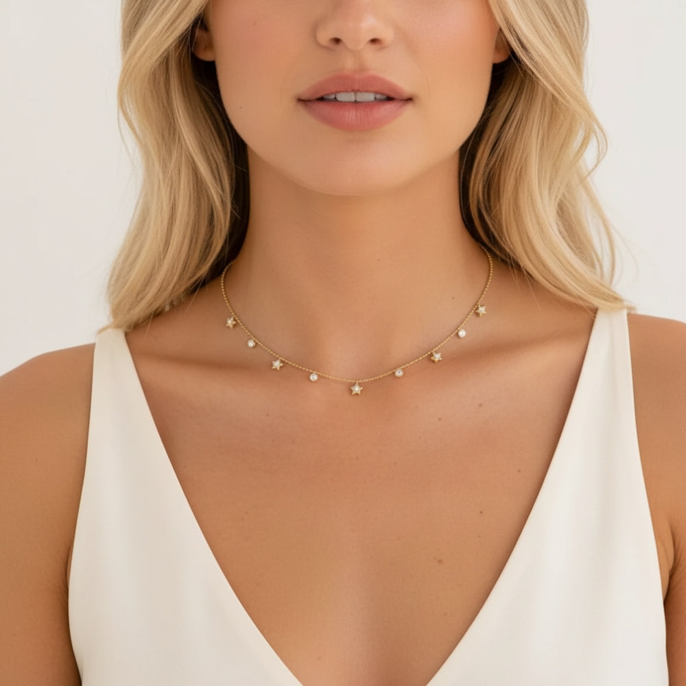A close-up of a blonde woman wearing a delicate gold chain necklace adorned with small star and crystal -like beads, paired with a white sleeveless top.