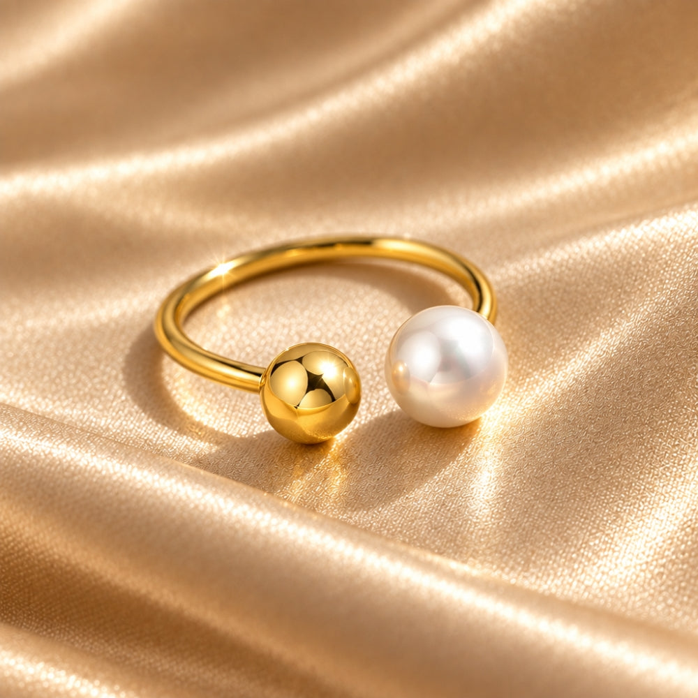 A gold-toned ring with one polished gold sphere and one white pearl bead, displayed on a draped champagne satin fabric background.