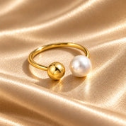 A gold-toned ring with one polished gold sphere and one white pearl bead, displayed on a draped champagne satin fabric background.