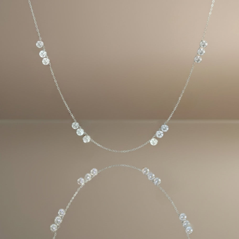 A delicate silver chain necklace featuring a series of small, clear faceted gemstones spaced along the chain, displayed against a warm beige background with a soft reflection below.
