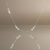 A delicate silver chain necklace featuring a series of small, clear faceted gemstones spaced along the chain, displayed against a warm beige background with a soft reflection below.