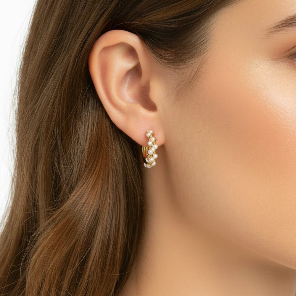 A close-up of a woman's ear wearing a small gold huggie hoop earring adorned with white pearls, set against her brown hair and skin.