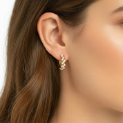 A close-up of a woman's ear wearing a small gold huggie hoop earring adorned with white pearls, set against her brown hair and skin.