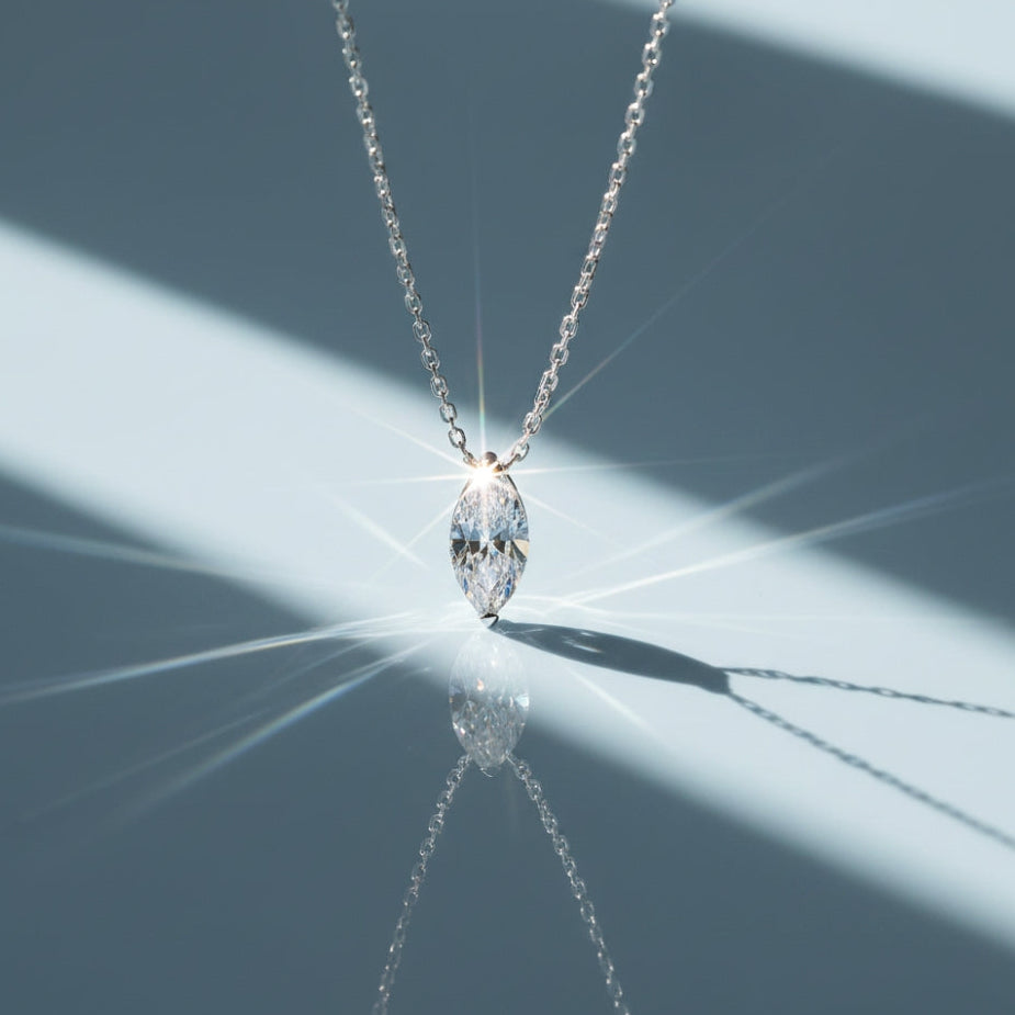 A silver chain necklace with a marquise-cut clear gemstone pendant, photographed on a reflective surface with dramatic diagonal light beams and a sharp reflection.