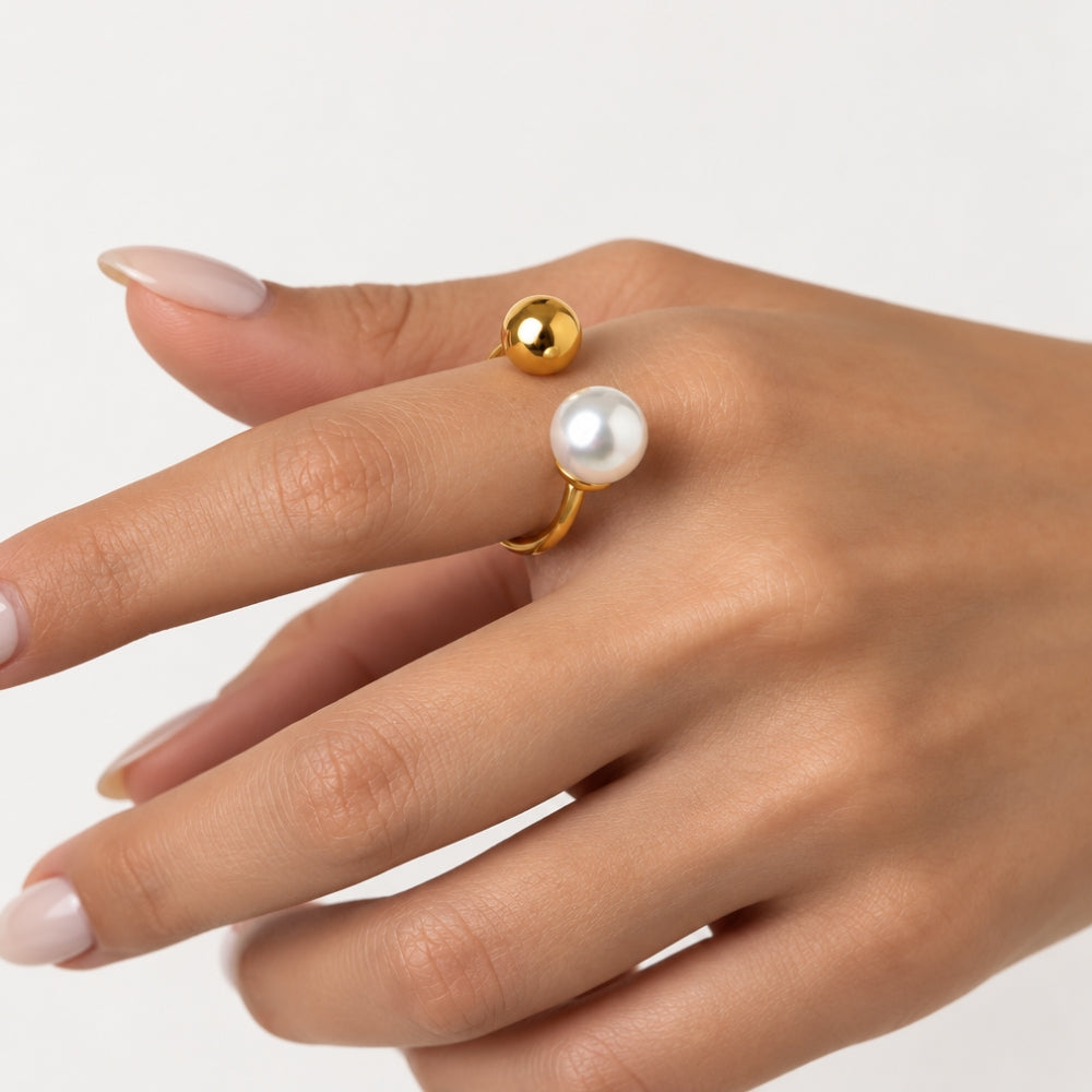 A gold ring with a white pearl and a small gold bead, shown worn on a hand against a light gray background.