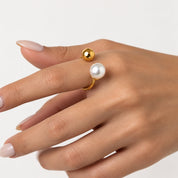 A gold ring with a white pearl and a small gold bead, shown worn on a hand against a light gray background.
