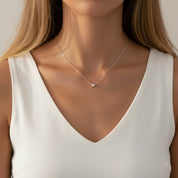 A close-up of a woman wearing a delicate silver chain necklace with a small, minimalist square pendant, styled with a white sleeveless V-neck top.