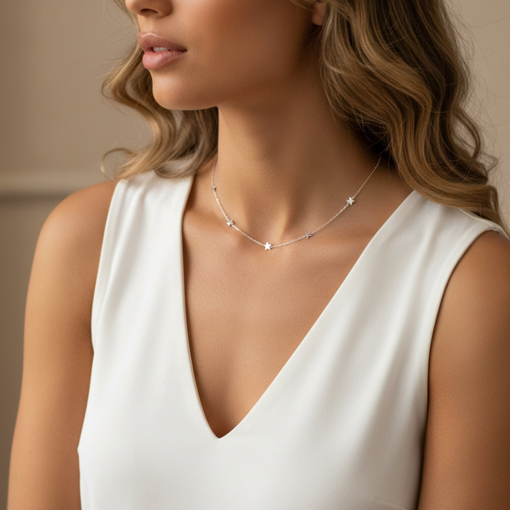 A close-up lifestyle shot of a woman wearing a delicate silver necklace with small star-shaped charms, paired with a white sleeveless V-neck top.