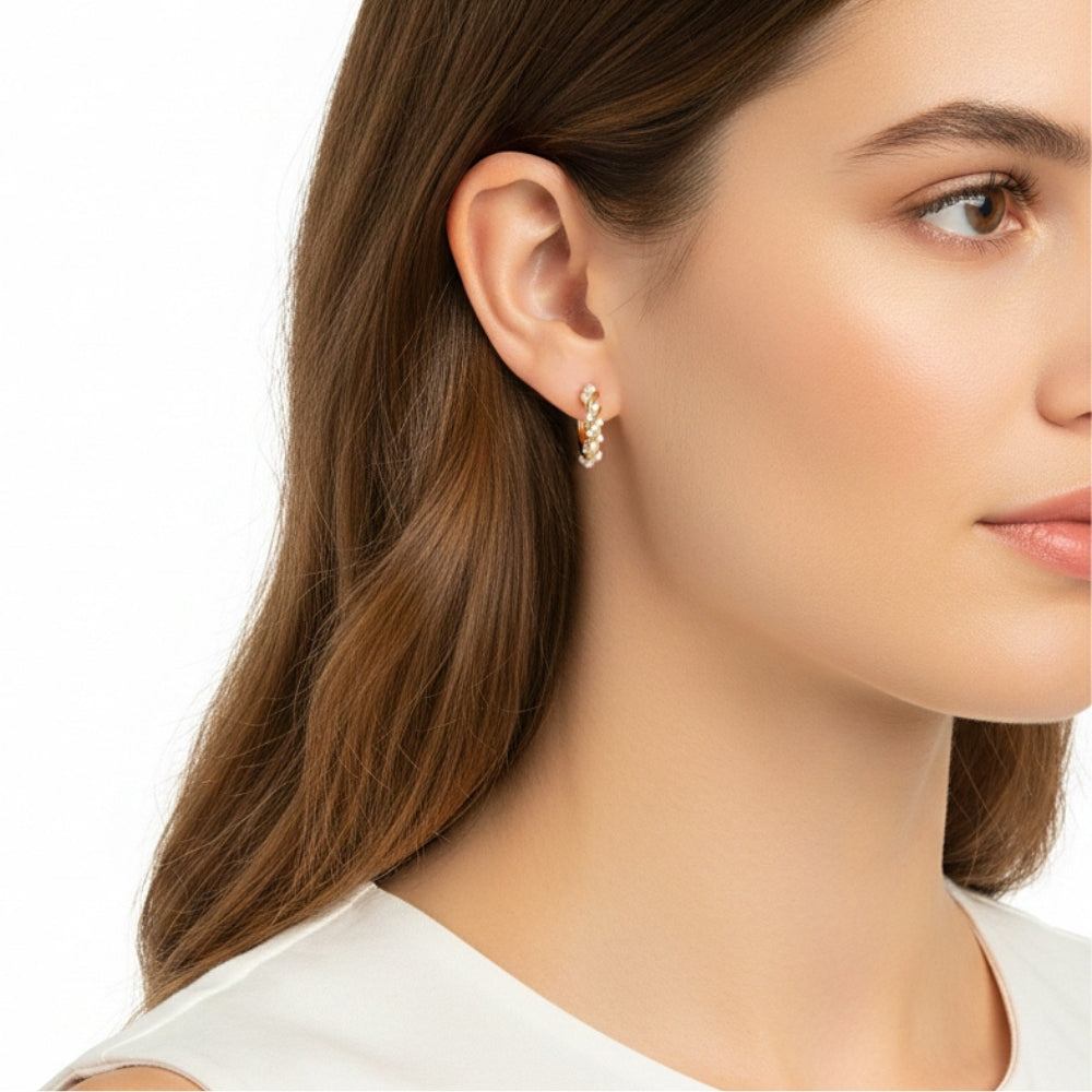 A close-up of a woman's ear wearing a delicate gold huggie hoop earring with small clear stones, shown against a clean white background.
