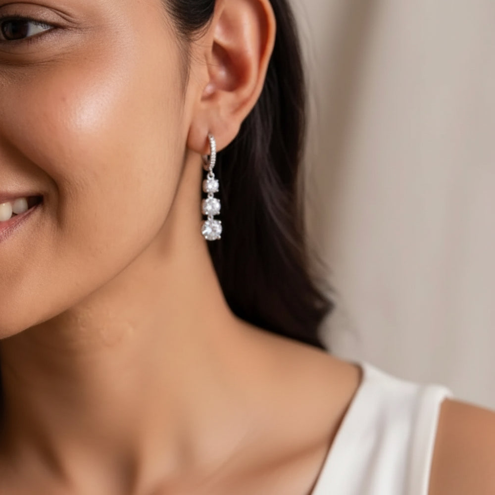 A close-up lifestyle shot of a woman wearing a silver drop earring with three clear round-cut gemstones. The earring is shown on her ear, with her face and shoulder visible against a soft, neutral background.