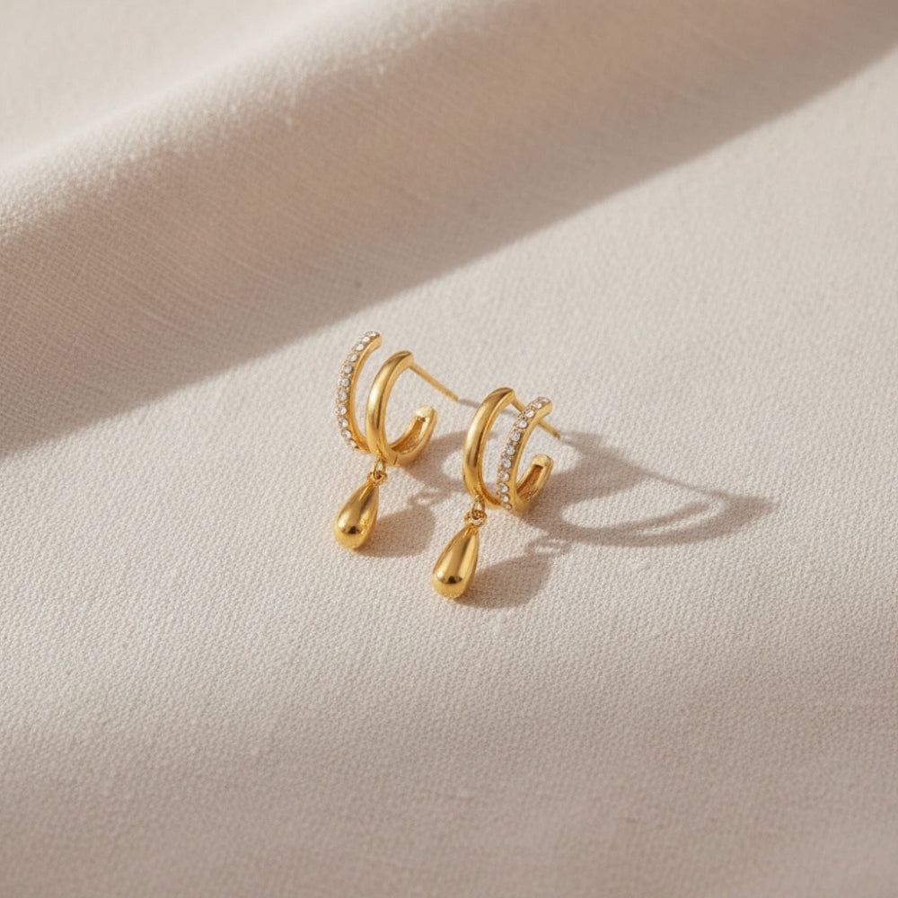 A pair of gold hoop earrings with teardrop pendants and small clear stone accents, displayed on a textured beige fabric background.