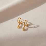 A pair of gold hoop earrings with teardrop pendants and small clear stone accents, displayed on a textured beige fabric background.