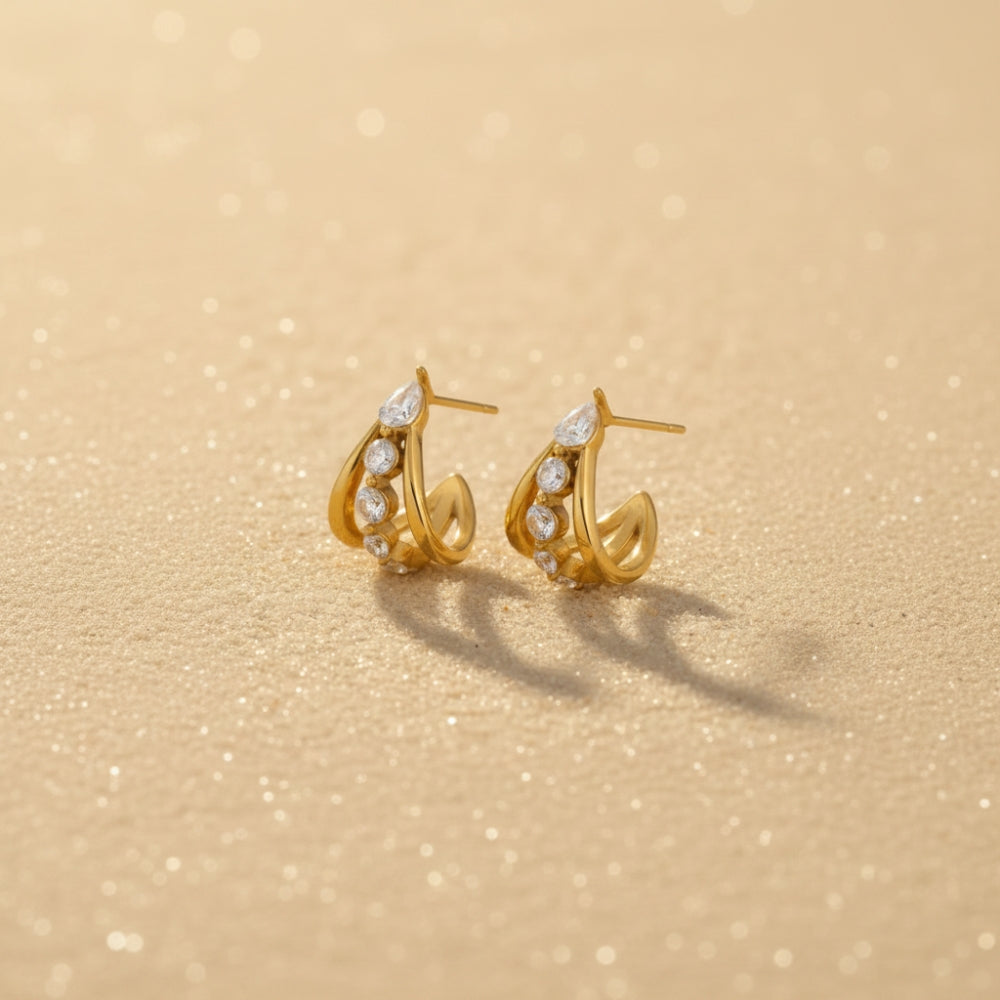 A pair of gold crescent-shaped hoop earrings with small clear stones, displayed on a textured beige surface with soft shadows.