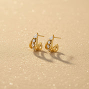 A pair of gold crescent-shaped hoop earrings with small clear stones, displayed on a textured beige surface with soft shadows.