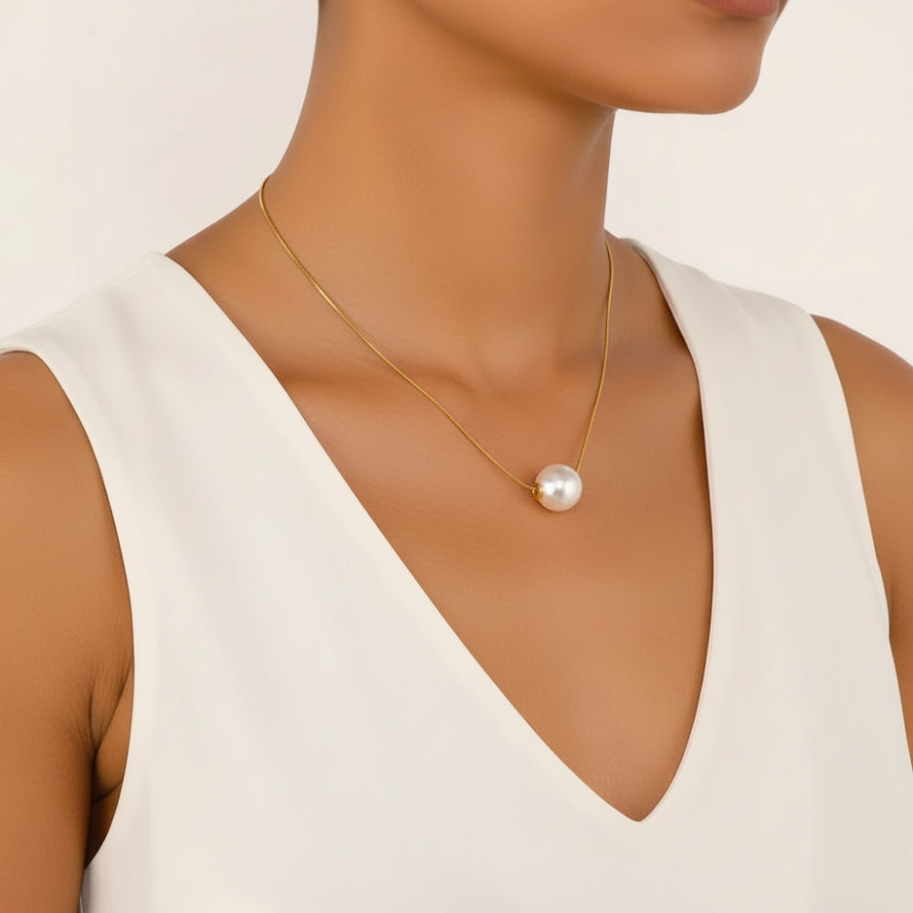 A close-up of a model wearing a delicate gold chain necklace with a single white pearl pendant, styled with a white sleeveless top against a clean off-white background.