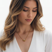 A woman with wavy blonde-highlighted hair wearing a delicate gold necklace with a small flower-like pendant, dressed in a white V-neck top against a clean white background.