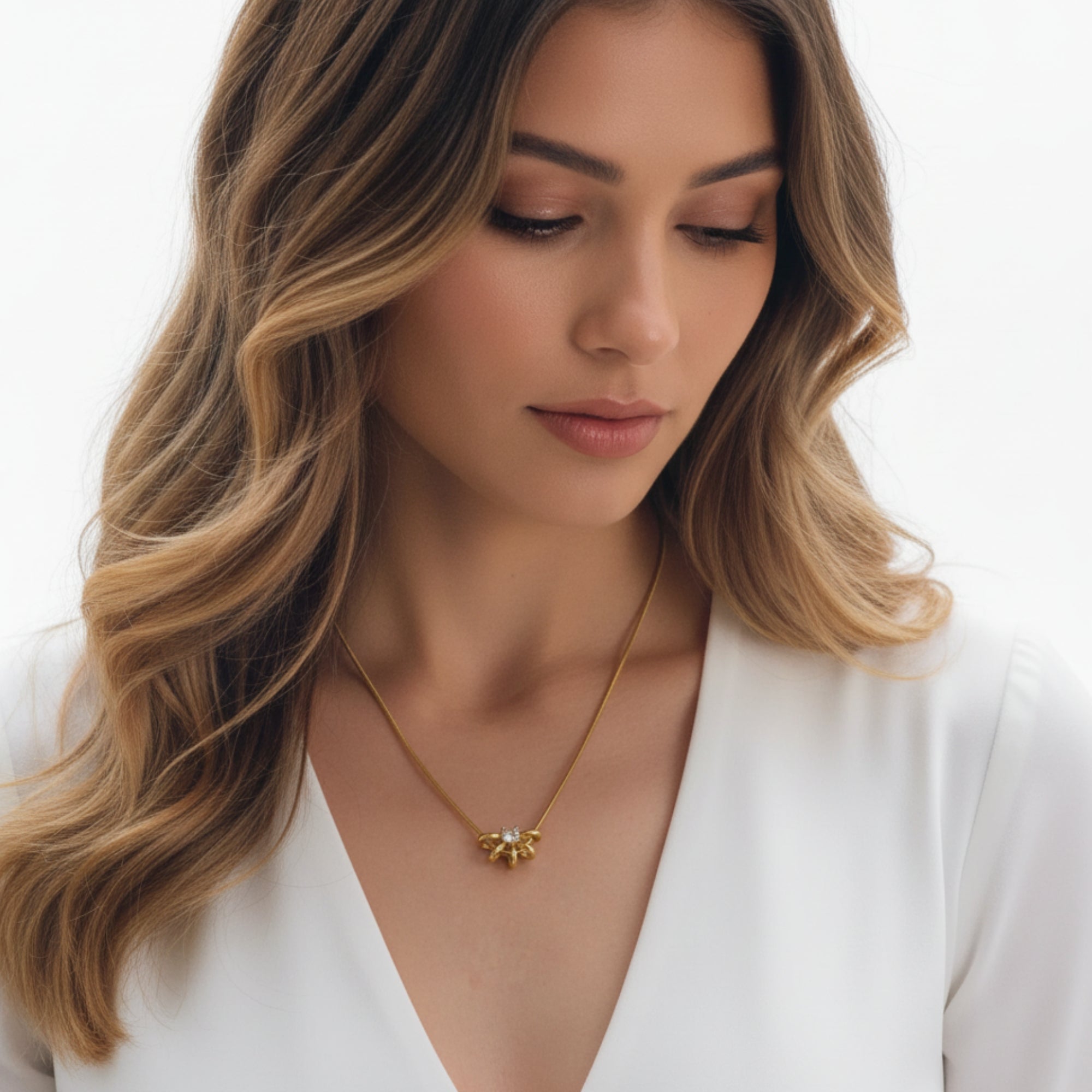 A woman with wavy blonde-highlighted hair wearing a delicate gold necklace with a small flower-like pendant, dressed in a white V-neck top against a clean white background.