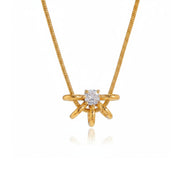 A gold necklace with a starburst pendant featuring a central round-cut clear gemstone, displayed on a clean white background with a soft reflection below.