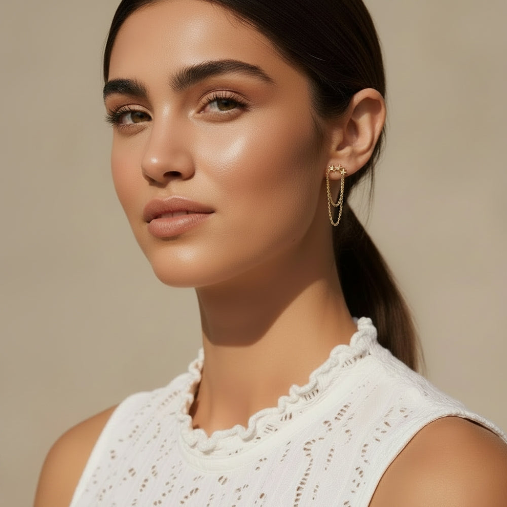 A close-up portrait of a woman with dark hair tied back, wearing a white textured sleeveless top and a delicate gold chain earring, set against a warm beige background.