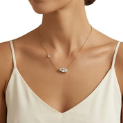 A close-up of a model wearing a delicate gold chain necklace with a teardrop-shaped clear crystal pendant and a small round clear crystal accent, paired with a cream-colored spaghetti strap top against a white background.