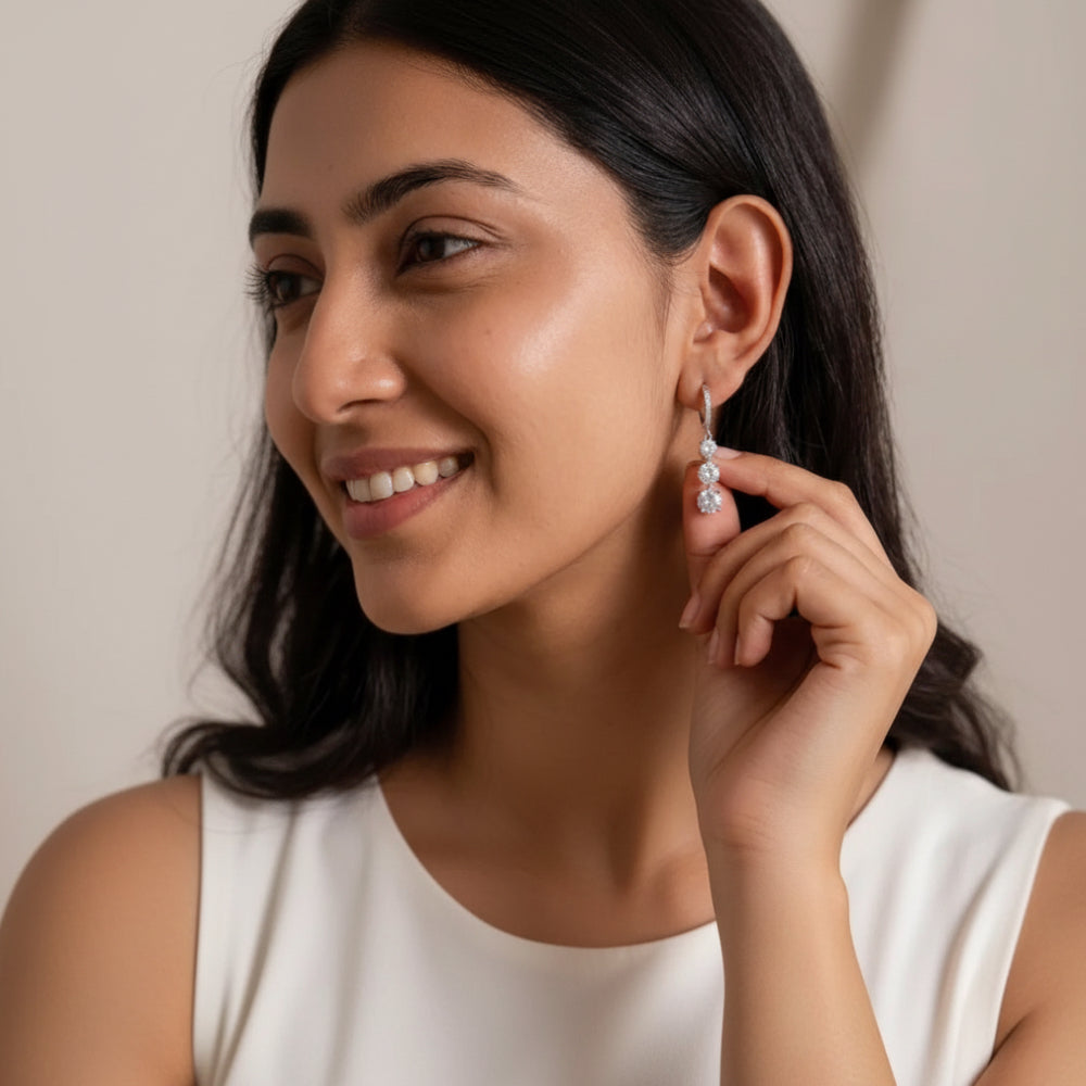 A woman wearing a delicate silver-toned dangle earring with small clear stones, shown in a close-up portrait against a soft beige background.
