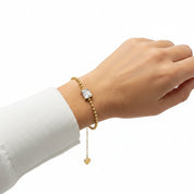 A gold beaded bracelet with a rectangular clear stone centerpiece and a dangling gold heart charm, shown worn on a person's wrist against a white background.