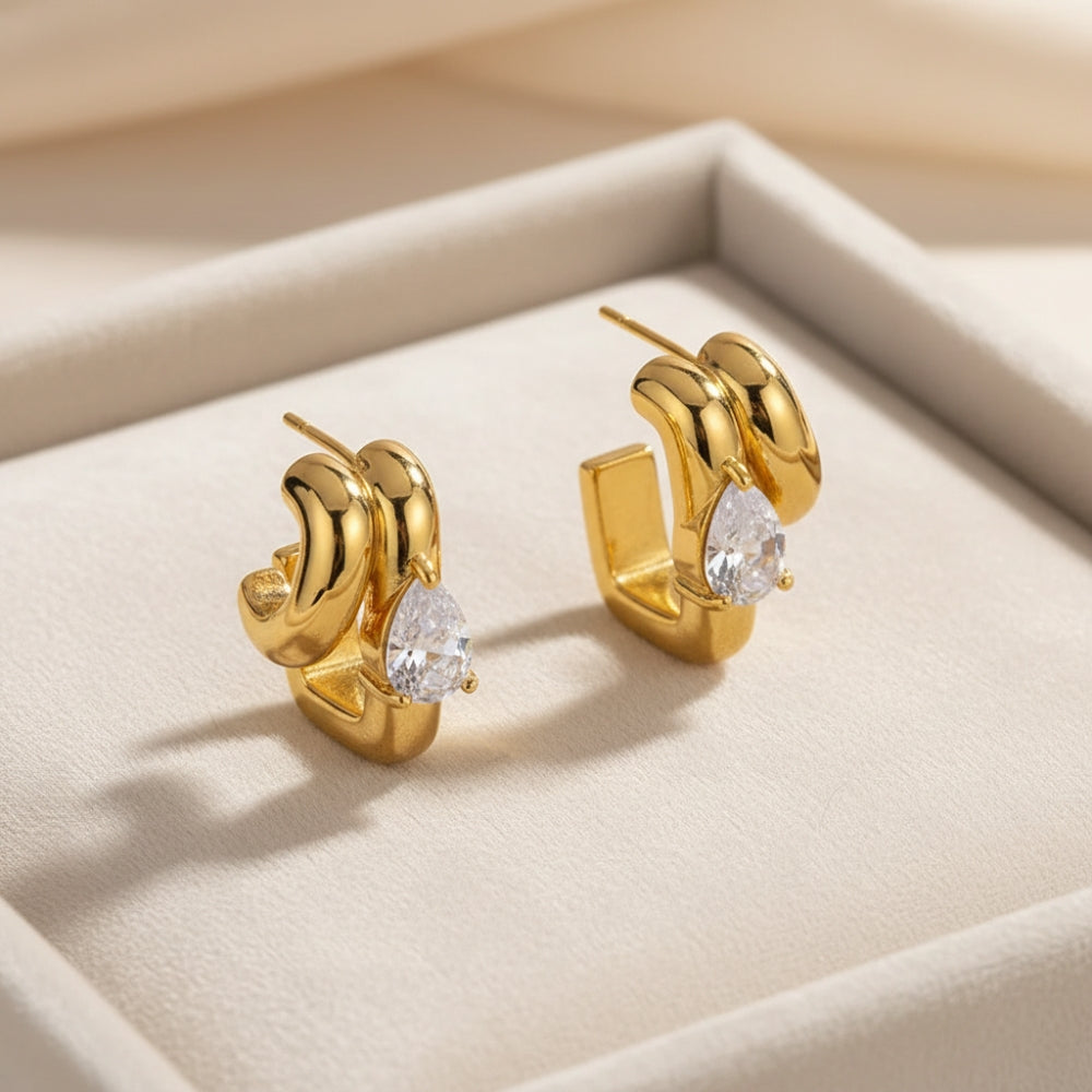 A pair of gold huggie hoop earrings with a teardrop-shaped clear gemstone, displayed on a beige jewelry box.