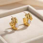 A pair of gold huggie hoop earrings with a teardrop-shaped clear gemstone, displayed on a beige jewelry box.