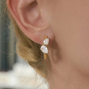 A close-up of a person's ear wearing a gold huggie hoop earring with two pear-cut clear gemstones.