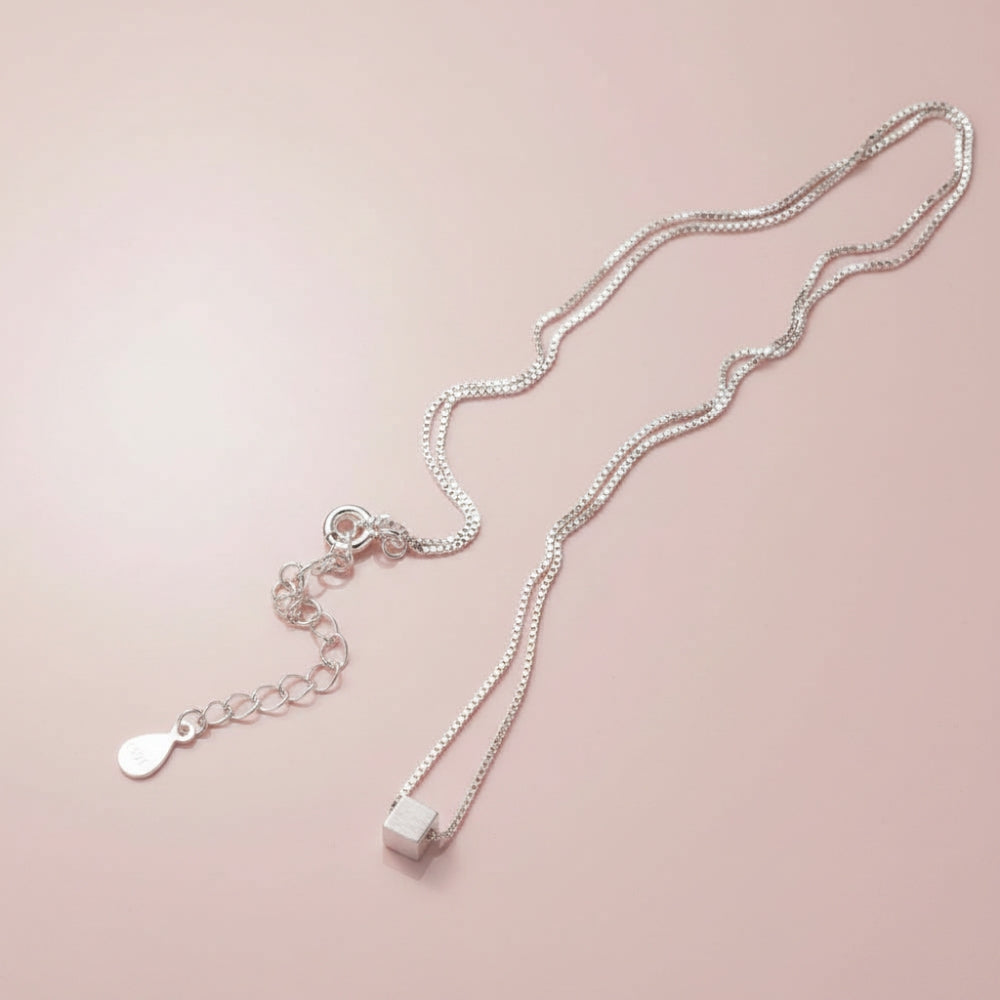 A delicate silver chain necklace with a small cube pendant and an adjustable chain extension featuring a teardrop-shaped charm, displayed on a soft pink background.