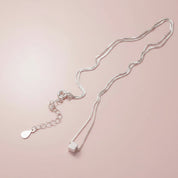 A delicate silver chain necklace with a small cube pendant and an adjustable chain extension featuring a teardrop-shaped charm, displayed on a soft pink background.