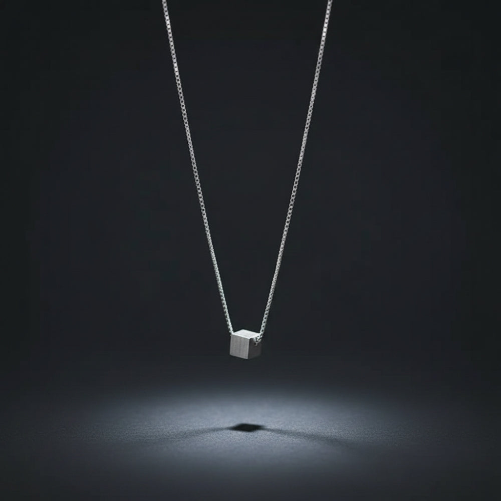 A silver cube pendant necklace suspended from a delicate silver chain, centered against a dark gray background with a spotlight effect below the pendant.
