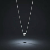 A silver cube pendant necklace suspended from a delicate silver chain, centered against a dark gray background with a spotlight effect below the pendant.