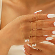 A close-up of a person's hand with white manicured nails, wearing a delicate gold necklace with a bar-shaped setting and three small white pearls, against a warm skin-toned background.