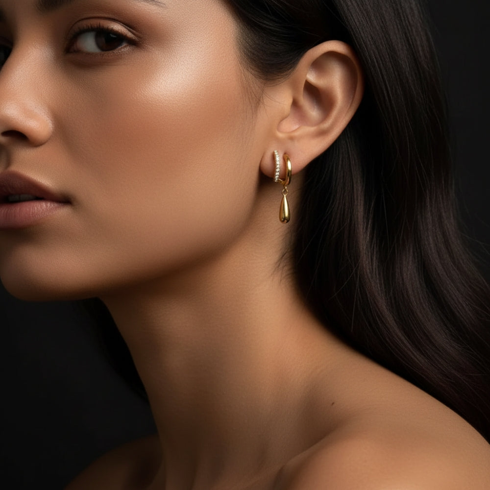 A close-up of a woman wearing a delicate gold huggie hoop earring with a small vertical row of clear stones and a teardrop-shaped gold drop pendant, set against a dark background.