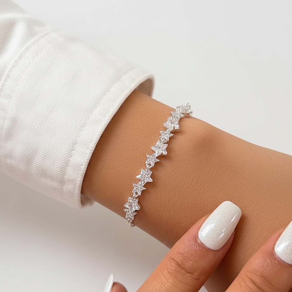 A delicate silver bracelet with a row of small star-shaped clear crystals, shown worn on a person's wrist. The person is wearing a white long-sleeve top, and their manicured nails are visible in the foreground against a clean white background.