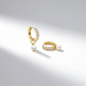 A pair of gold huggie hoop earrings, one dangling with a small white pearl and the other resting flat with a small white pearl and a row of clear pavé stones, set against a clean white background with soft shadows.