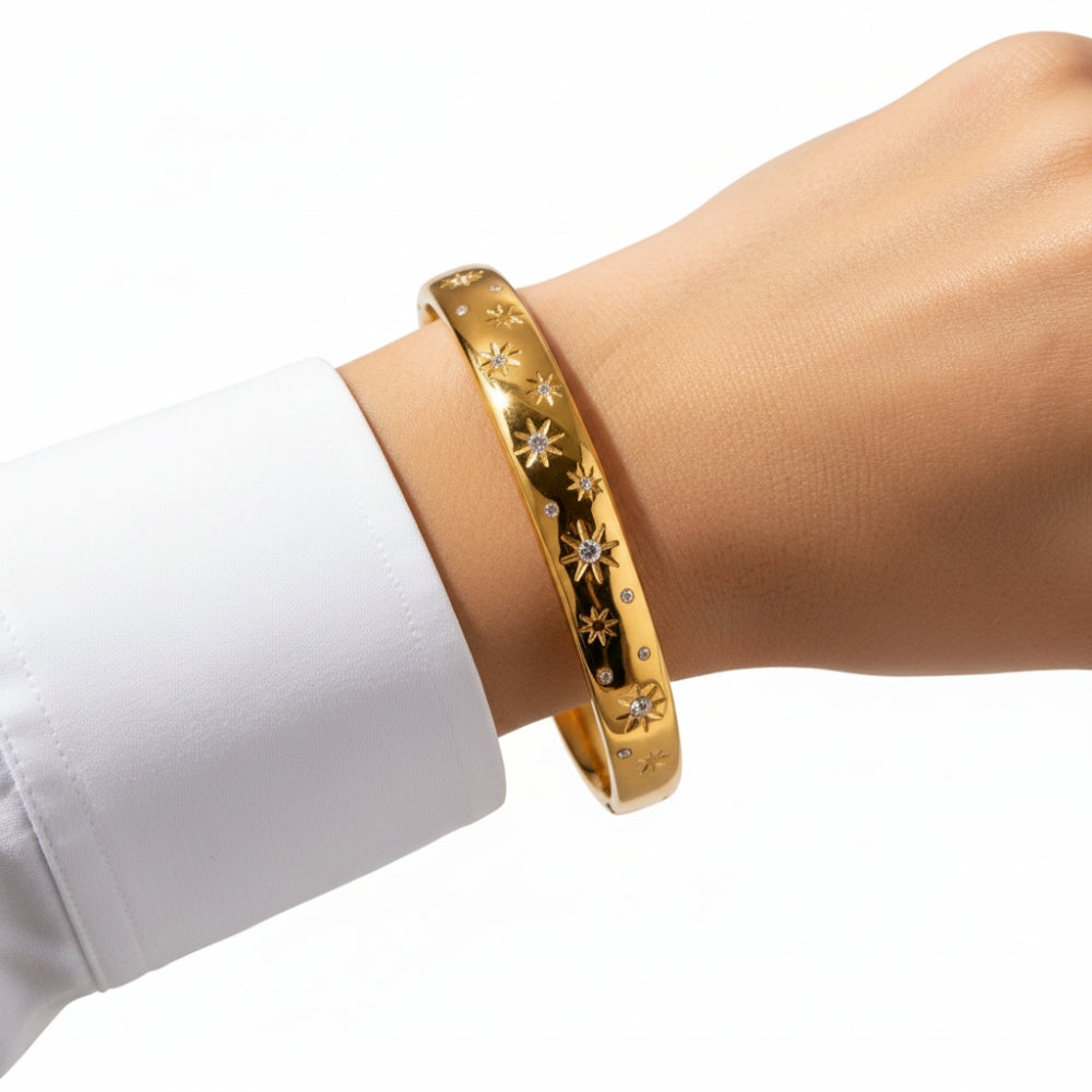 A gold bangle bracelet with star and diamond-like engravings, shown worn on a person's wrist with a white dress shirt cuff visible.