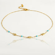 A delicate gold chain necklace featuring small gold star charms, tiny turquoise blue beads, and clear crystal accents, displayed on a clean white reflective surface.