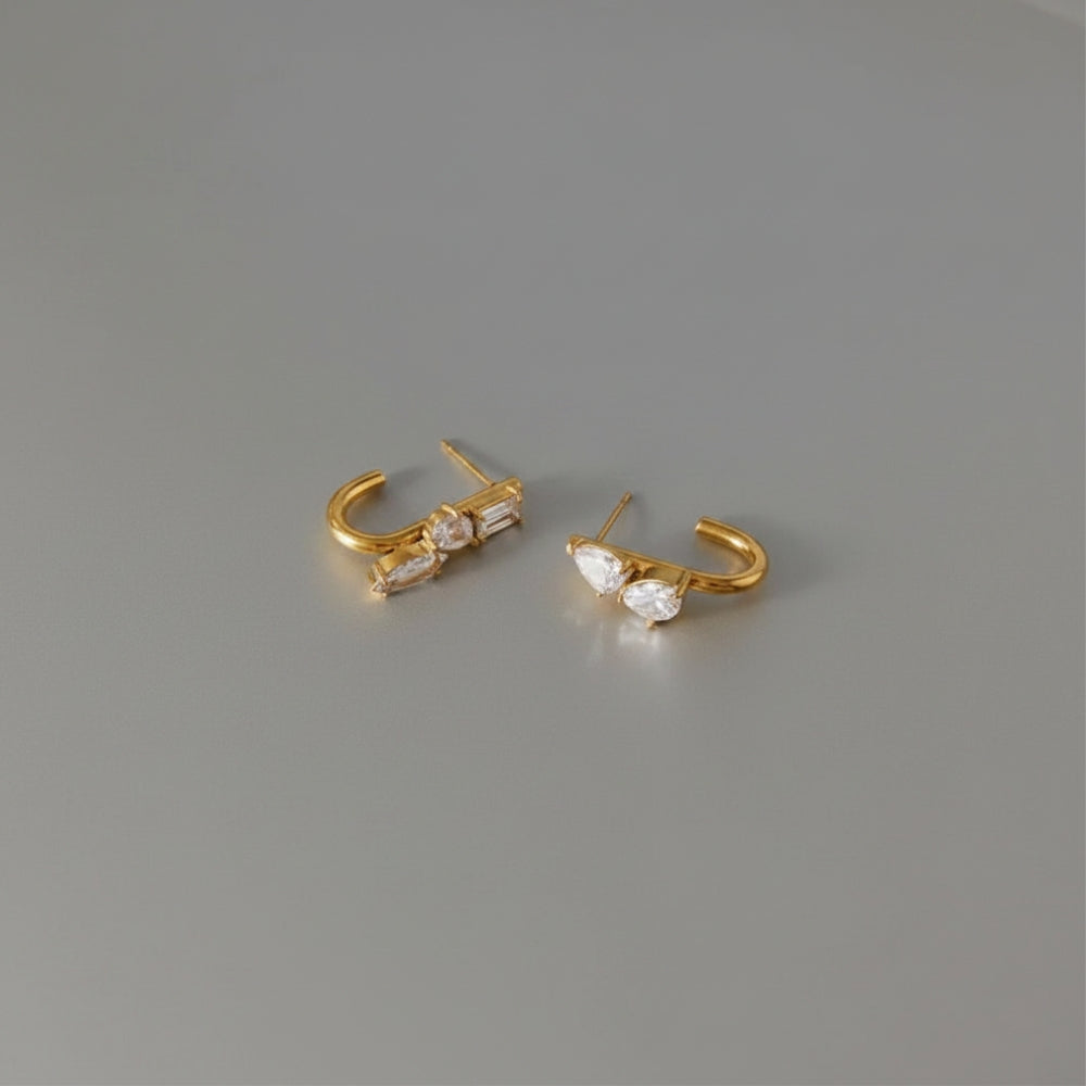 A pair of gold hoop earrings with clear crystals, displayed on a neutral gray background.