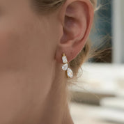A close-up of a model's ear wearing a gold dangle earring with three clear rectangular and oval gemstones.