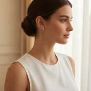 A woman in a white sleeveless top wearing a small gold hoop earring, shown in profile against a soft beige indoor background.