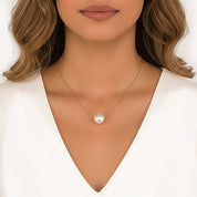 A close-up of a woman wearing a delicate gold chain necklace with a single lustrous white pearl pendant, styled with a white V-neck top against a clean white background.