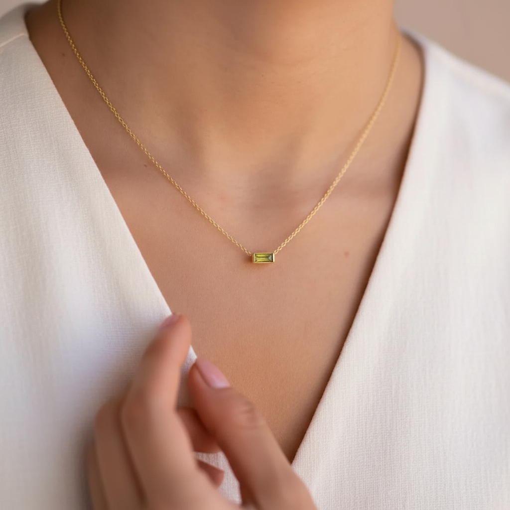 A close-up of a model wearing a delicate gold chain necklace with a small rectangular olive  green gemstone pendant, styled with a white V-neck top.
