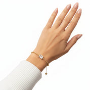A gold beaded bracelet with a rectangular clear gemstone and a small dangling heart charm, shown worn on a woman's wrist against a white background.
