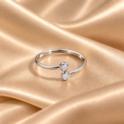 A silver open ring with two small round clear gemstones, displayed on draped champagne-colored satin fabric.