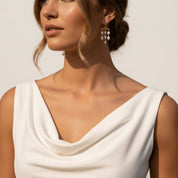 A close-up of a woman wearing gold dangle earrings with white pearl-like beads, paired with a white draped sleeveless top against a warm off-white background.